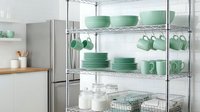 Chrome wire shelving unit in a modern kitchen displaying mint green ceramic dishes, bowls, mugs, and glass jars for organized storage.