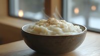 Creamy mashed potatoes with melted butter and fresh ground pepper served in rustic brown ceramic bowl on wooden table, soft window lighting creates cozy atmosphere.