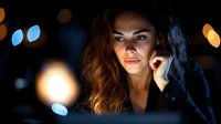 Young woman with wavy hair illuminated by computer screen in dark room with bokeh lights in background, creating dramatic lighting on face.