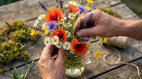 Florist arranging vibrant wildflowers including red gerberas, purple lavender, and white daisies in glass vase outdoors. Gardening, floral design, and nature arrangement concept.