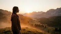 Woman in brown jacket standing on mountain overlooking misty valley at sunrise, contemplating scenic alpine landscape with forested peaks and golden light.