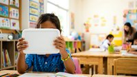 Young girl with dark hair and medium skin tone holding a tablet device in front of her face in a colorful classroom setting with educational materials and other students in background.