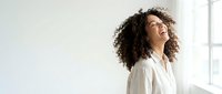 Happy young African American woman with curly hair laughing joyfully in bright white shirt by window. Natural light portrait expressing pure happiness and positivity.