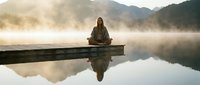 Young woman meditating in lotus position on wooden dock over misty lake at sunrise with mountain reflections creating peaceful zen atmosphere for wellness.