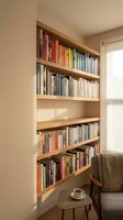 Built-in wooden bookshelf filled with colorful books in a bright cozy reading nook with armchair and tea cup by sunny window.