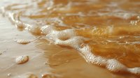 Gentle ocean wave with white foam washing onto sandy beach at sunset, creating natural patterns and textures in golden evening light.