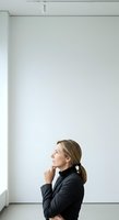 Middle-aged woman with shoulder-length brown hair, Caucasian ethnicity, wearing black blazer, standing in modern minimalist interior with white walls, looking upward thoughtfully, professional business portrait.