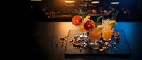 Craft cocktails with blood orange garnish on slate board in upscale bar setting with warm ambient lighting and blurred background for hospitality design.