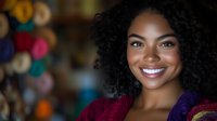 Young African American woman with curly hair smiling warmly in colorful traditional market setting with vibrant textiles and crafts in background.