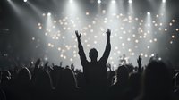 Silhouette of person with raised arms at music concert against bright stage lights and bokeh effect, crowd enjoying live performance in dark atmosphere.