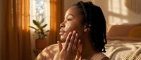 Young African American woman applying skincare cream in sunny bedroom during morning routine. Natural beauty and self-care wellness lifestyle concept.