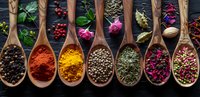 Aromatic spices and dried flowers in wooden spoons on dark background, colorful cooking ingredients including turmeric, paprika, peppercorns and rose petals for culinary design.