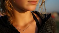 Close-up portrait of young woman with wavy hair wearing delicate necklace at golden hour, soft natural lighting creating warm glow on skin.