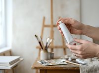 Close-up of hands holding white spray bottle in bright studio workspace with art supplies and easel, suitable for product mockups and creative professional settings.