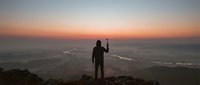 Silhouette of hiker with raised arm celebrating success on mountain peak during golden sunset over ocean waves and coastline below.