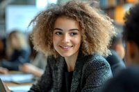 Young mixed race woman with natural curly afro hair smiling confidently in office environment wearing casual grey sweater, professional workplace portrait.