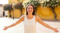 Woman with long dark hair, Hispanic ethnicity, wearing a white sleeveless tank top, smiling with arms outstretched in a welcoming gesture against a yellow colonial building backdrop. Suitable for lifestyle, travel, and happiness concepts.