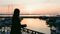 Young woman with dark hair, viewed from behind, standing at waterfront railing at sunset, overlooking marina with sailboats and fishing vessels, golden hour lighting, peaceful coastal scene.