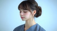Young Asian female healthcare worker with updo hairstyle and bangs wearing blue medical scrubs uniform against light background, eyes closed in contemplative expression.