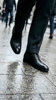 Businessman walking on wet city street in black leather dress shoes and dark suit pants during rush hour with blurred pedestrians in urban environment.