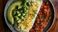 Fresh halibut fillet topped with eggs, served alongside diced avocado, cucumber and pico de gallo salsa in gray ceramic bowl on wooden surface.