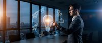 Professional businessman in suit holding glowing lightbulb with digital innovation icons in modern office overlooking city skyline at sunset.