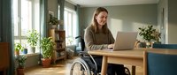 Man in wheelchair working at wooden desk with laptop in bright home office with plants and natural light from windows.