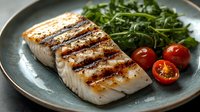 Grilled white fish fillet with charred marks served on blue ceramic plate alongside fresh arugula salad and halved cherry tomatoes, close up view of seafood dinner.