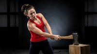 Athletic Caucasian woman in red tank top swinging sledgehammer at wooden block in gym setting. Functional fitness training workout exercise routine.