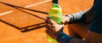 Athlete holding sports drink bottle on clay tennis court during match break, staying hydrated and refreshed.