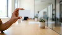 Hand holding blank white business card in modern office workspace with wooden desk and glass partition walls, suitable for corporate branding and professional networking materials.