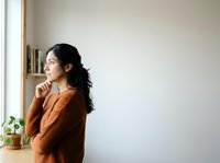 Young East Asian woman in rust-colored knit sweater standing thoughtfully by wooden desk with potted plant, looking away contemplatively in bright minimalist interior space.