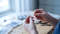 Hands threading needle for embroidery or sewing project on wooden table near window with natural light creating peaceful crafting atmosphere.