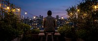 Man sitting on rooftop overlooking illuminated city skyline at dusk, surrounded by potted plants and urban lights, contemplative evening scene for lifestyle and urban living concepts.