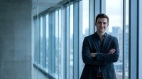 Confident young Caucasian businessman with crossed arms standing in modern office corridor with large windows overlooking city skyline for corporate success.