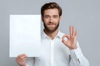 Young bearded man in white shirt showing okay gesture while holding blank paper mockup against gray background. Perfect for advertising or presentation.