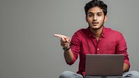 Young South Asian man in burgundy shirt pointing while sitting cross-legged with laptop against gray background, expressing explanation or instruction.