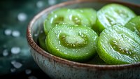 Sliced green kiwi fruit in ceramic bowl with water droplets, showcasing juicy texture and seeds on dark background.