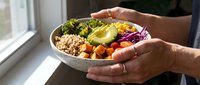 Person holding a nutritious bowl with quinoa, roasted vegetables, avocado and purple cabbage by a window for healthy eating and meal prep content.