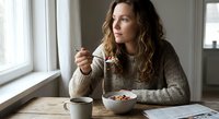 Young Caucasian woman in cozy sweater enjoying healthy breakfast bowl at home kitchen table by window with natural morning light and coffee cup.