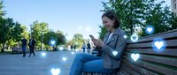 Young woman using smartphone on park bench with floating heart icons representing social media engagement and digital connection in urban setting.