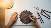 Adult coloring mandala art therapy session with hands drawing intricate geometric pattern using colored pencils for stress relief and mindfulness practice.