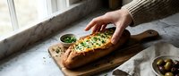 Woman in cozy sweater preparing fresh herb-topped bread on wooden cutting board by window. Homemade cooking and artisanal food preparation concept.