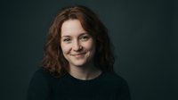 Young Caucasian woman with curly brown hair smiling warmly at camera wearing dark sweater against moody background for professional headshot portraits.