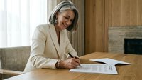 Mature Caucasian businesswoman with gray hair signing documents at wooden desk in modern office, professional executive reviewing contracts.