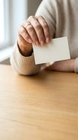 Middle-aged man with medium skin tone wearing beige sweater holding blank white card on wooden table, suitable for business card mockups and corporate branding.