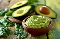 Fresh guacamole dip in rustic wooden bowl with ripe avocado halves and green spinach leaves on wooden table, selective focus on creamy avocado paste.