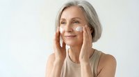 Mature Caucasian woman applying facial cream with gentle touch, showcasing anti-aging skincare routine and natural beauty confidence in minimalist setting.