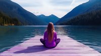 Young woman in purple attire sitting cross-legged on wooden pier overlooking serene mountain lake at dawn, practicing meditation and mindfulness in nature.