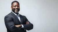 Confident African American businessman in formal suit with arms crossed, smiling against neutral gray background.